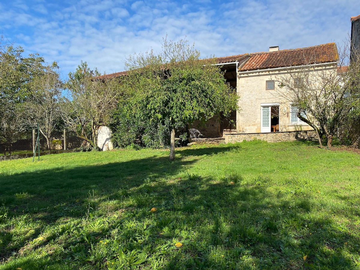 The exterior features a spacious garden with gently rolling grass and mature trees, providing ample shade. A charming stone house with a tiled roof is partially visible, along with an open area that allows for outdoor relaxation. The sky is clear with scattered clouds.