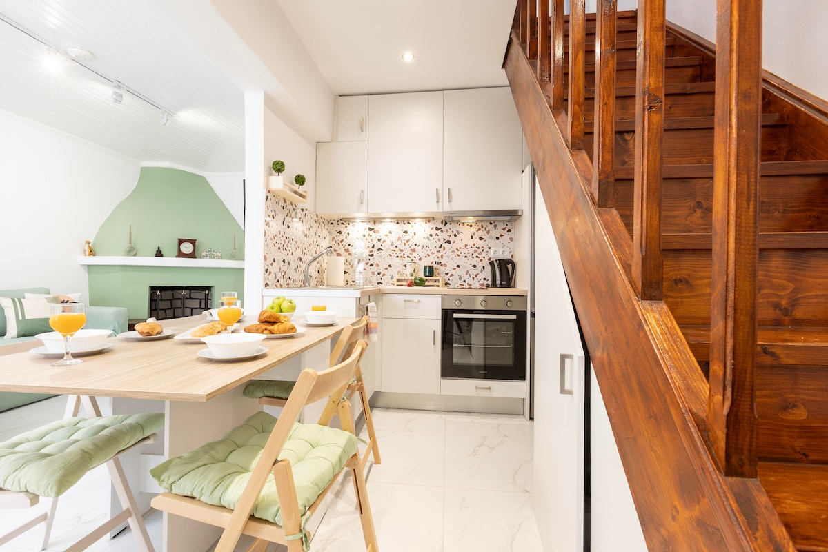 A modern kitchen and dining area is shared with a cozy living space. A wooden dining table is set with plates and drinks, while light green cushions adorn the dining chairs. The kitchen features integrated appliances and decorative tiles that add a touch of warmth.