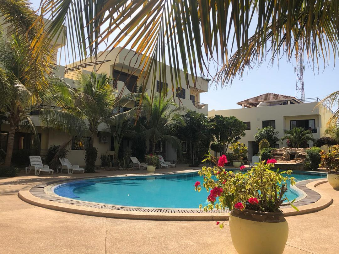 A landscaped pool area is surrounded by palm trees and colorful plants. The circular pool is visible in the foreground, with lounge chairs positioned around it. The surrounding buildings are multi-storied, showcasing balconies decorated with greenery.
