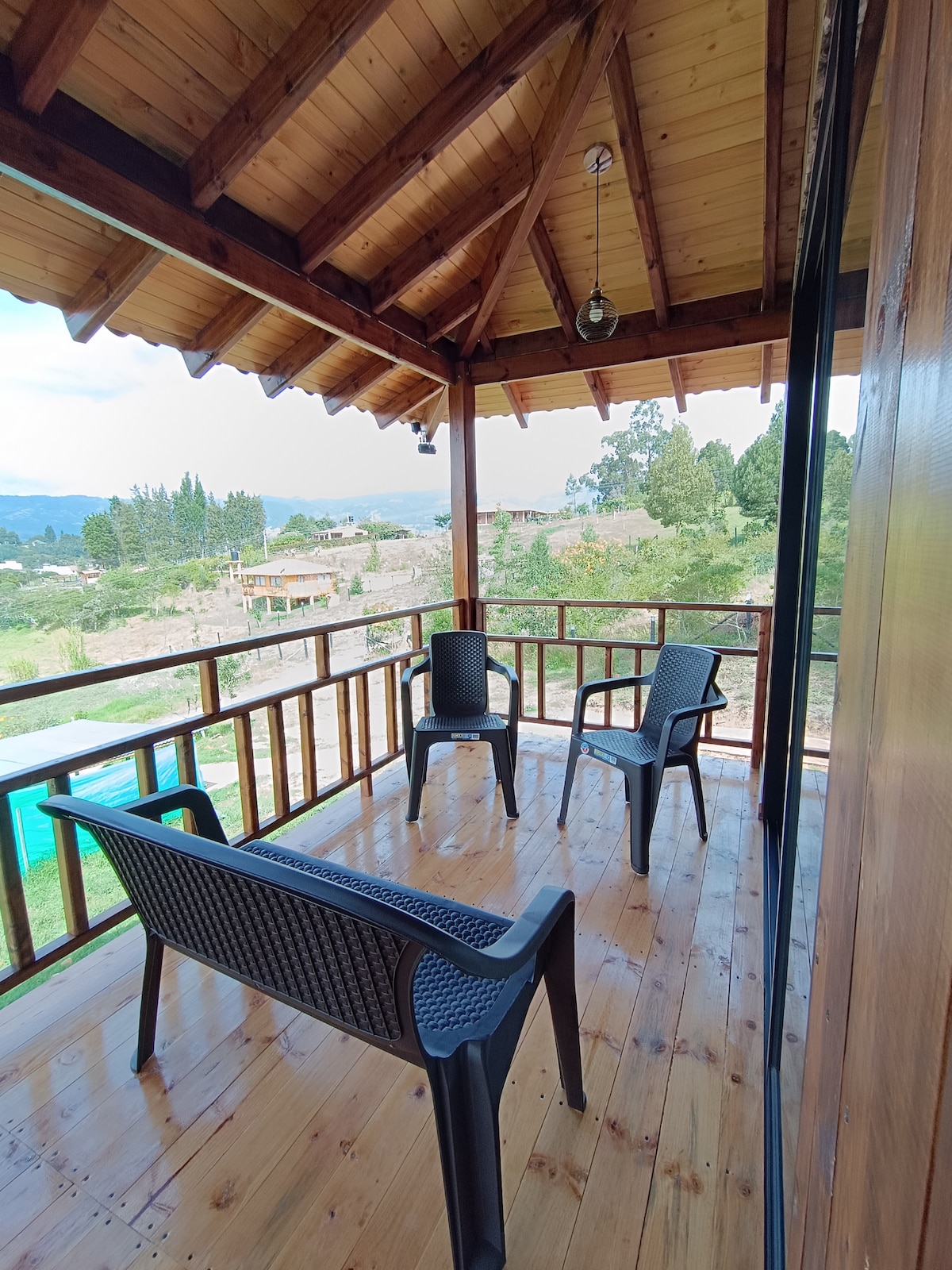 A spacious balcony is featured, showcasing a wooden roof with exposed beams. Two black chairs and a matching loveseat are arranged on the polished wooden floor, offering a serene view of the surrounding landscape and distant hills.