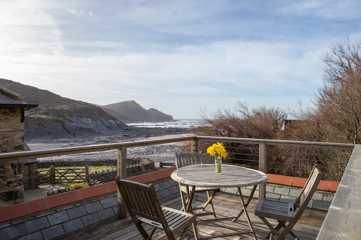 A wooden deck features a round table with a vase of yellow flowers at its center. Surrounding the table are four wooden chairs. In the background, expansive views of the beach and rolling hills are visible under a partly cloudy sky.