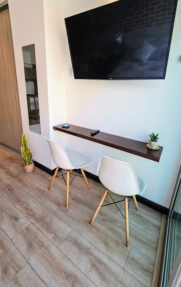 A sleek workspace area features two white chairs positioned beneath a mounted television. A minimalist shelf holds a small plant and a remote control, with a mirror reflecting light across the space. The light-colored wooden flooring contributes to a contemporary ambience.