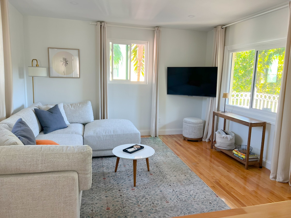 An open-concept living area features a light-colored sectional sofa with decorative pillows. A coffee table sits in front of the sofa, and a mounted television is positioned on the wall. Large windows allow natural light to fill the room, which is complemented by soft curtains.