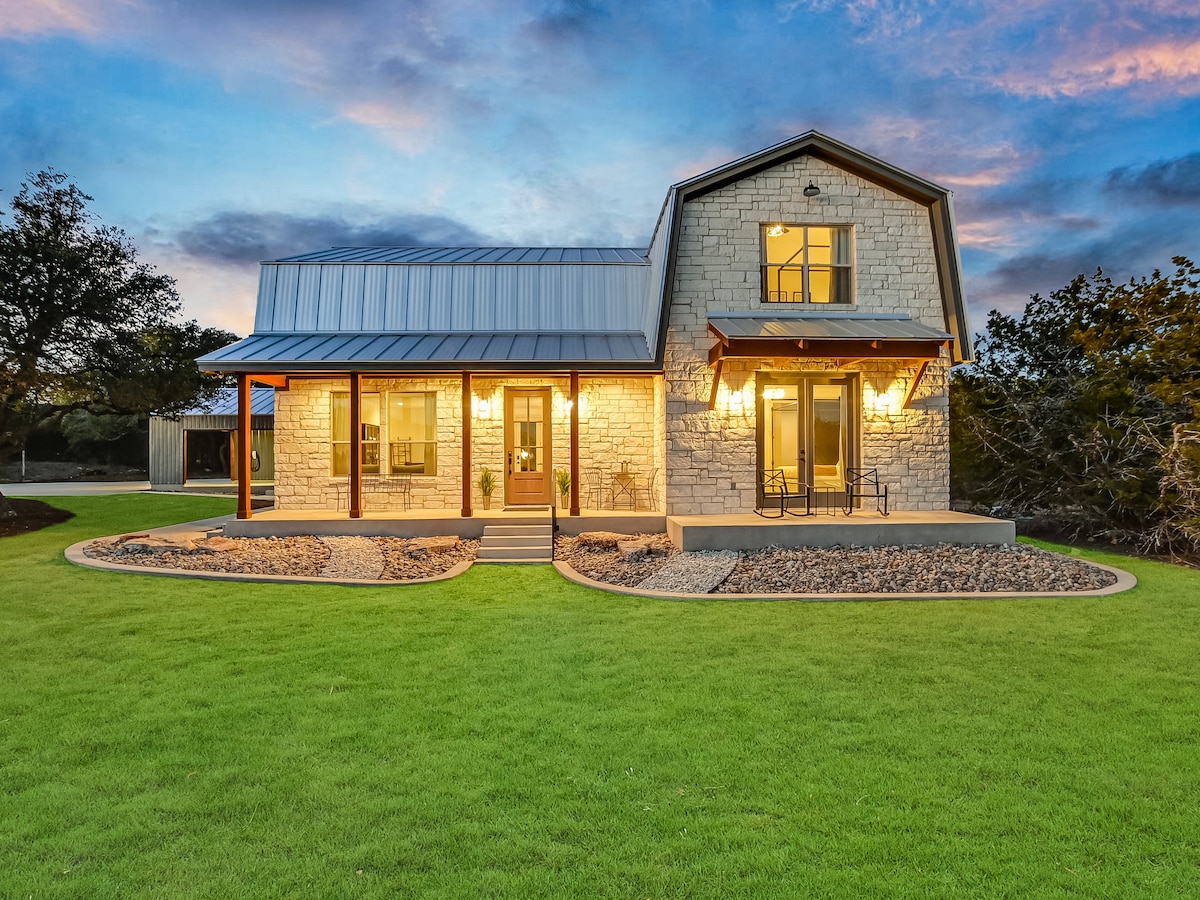 The exterior of the farmhouse is highlighted, featuring a combination of stone and metal siding. A welcoming porch with seating is visible, surrounded by well-maintained landscaping and open green space. The evening sky adds a soft glow, enhancing the inviting atmosphere of the home.