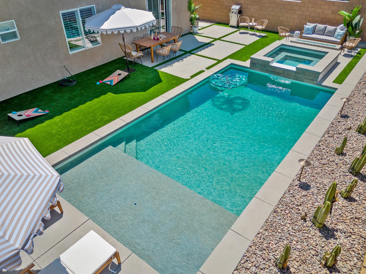 A private outdoor space features a heated swimming pool and a spa, surrounded by lush green grass and modern seating. Sun loungers sit beneath striped umbrellas, while a dining area with a table is visible nearby. Cornhole boards are placed on the grass, adding to the leisure setup.