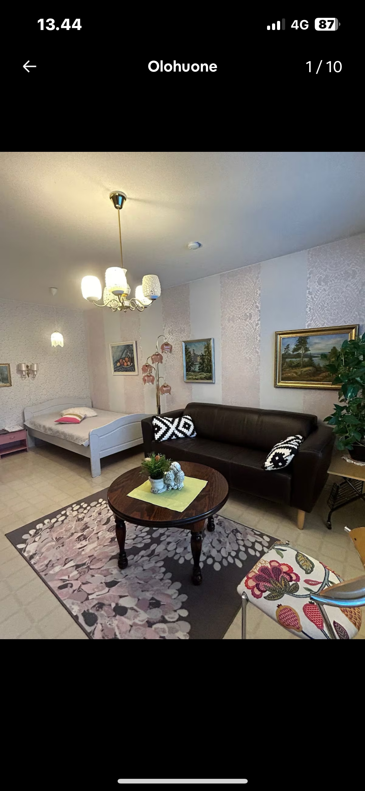 A living room area features a dark brown couch adorned with patterned cushions. A small round table sits in front, decorated with a flower arrangement and a colorful cloth. Light fixtures hang from the ceiling, and framed paintings are mounted on the walls, enhancing the space.
