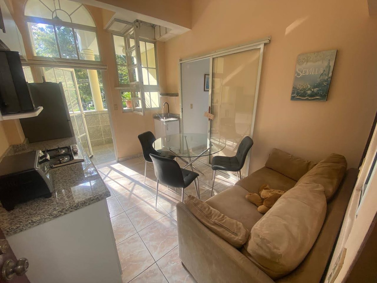 A comfortable living area is visible, featuring a spacious light-colored sofa and a glass dining table surrounded by four black chairs. Natural light filters in through large windows, complementing the warm tones of the walls and tile flooring.