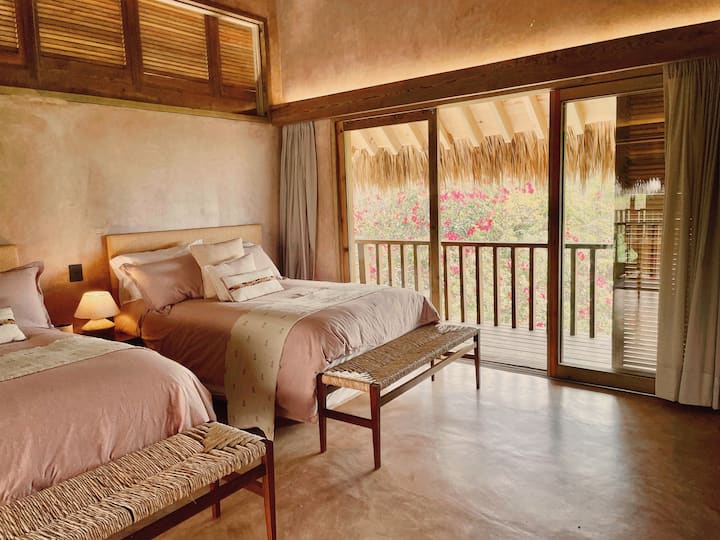- Acogedor dormitorio con camas dobles y vistas desde el balcón   - Destacan los tonos rosas suaves y las texturas tejidas   - Un refugio tranquilo rodeado de flores tropicales  