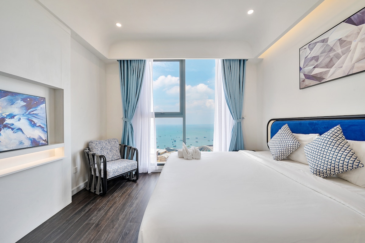 A bright bedroom is showcased with a spacious bed adorned with geometric patterned pillows. Large windows offer expansive views of the ocean and distant boats. A comfortable armchair sits in the corner, and soft blue curtains frame the view, creating a serene atmosphere.