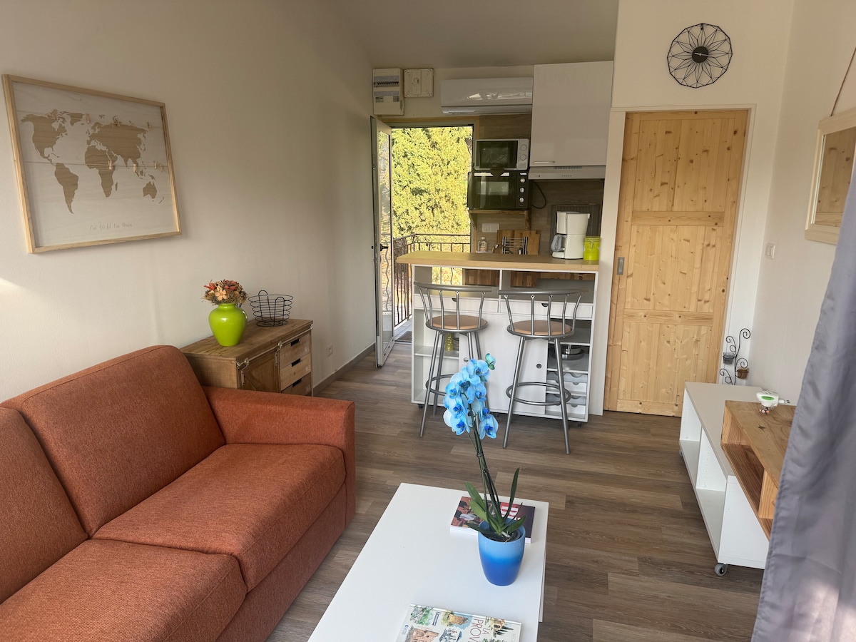 A bright living area is presented, featuring an orange sofa and a wooden coffee table. In the background, a small kitchenette is visible, accompanied by two bar stools at an elevated counter. A door opens to an outdoor view, with a decorative plant on the table.