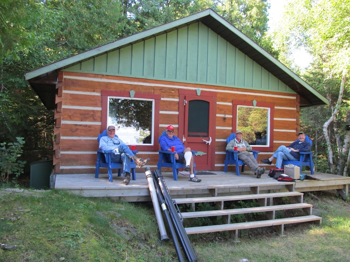 A rustic cabin is set amid greenery, featuring a wooden façade and a sloped roof. A porch with steps and blue chairs provides seating for guests, who are seen relaxing. The surrounding area includes trees and a lawn, creating a peaceful outdoor setting.