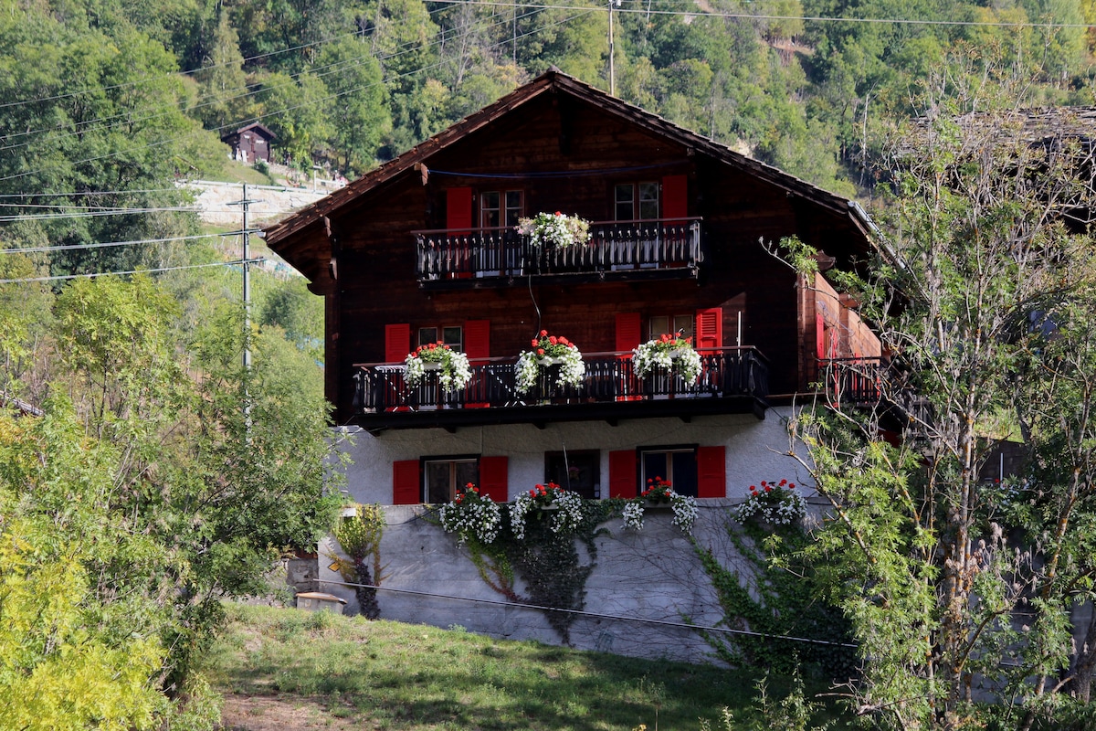 A charming wooden chalet is surrounded by lush greenery, showcasing vibrant flower boxes on each balcony. The red shutters add a pop of color to the traditional structure, which is set against a backdrop of rolling hills and trees.