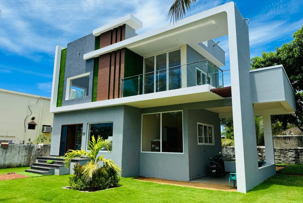 A modern two-story villa is showcased, with large glass windows allowing natural light to fill the interior. The exterior features a mix of grey and green walls, surrounded by lush greenery and a well-maintained lawn, creating an inviting and open atmosphere.