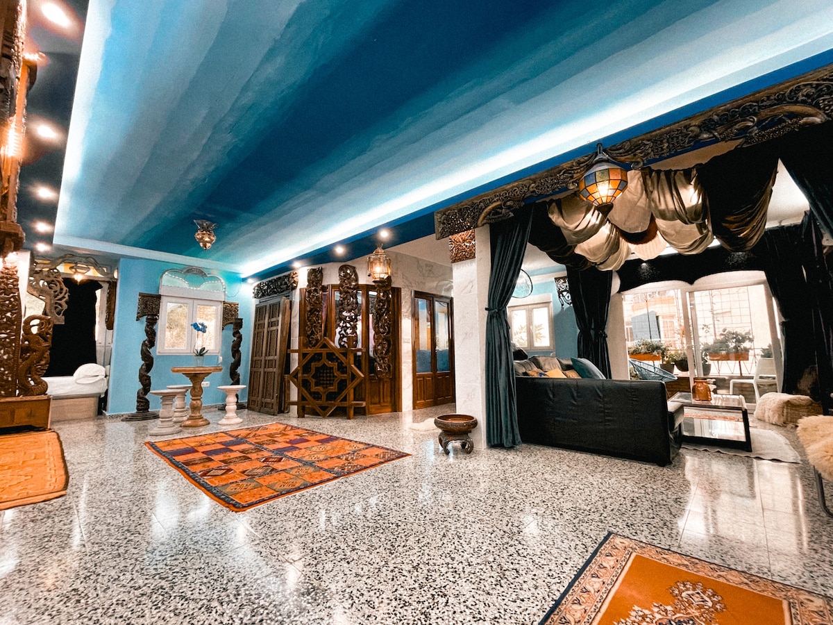 A spacious area is defined by intricate architectural details and artistic decorations. Elegant light fixtures hang from a beautifully painted ceiling. Decorative rugs add warmth to the polished floor, while plush seating invites relaxation in a cozy corner. Large windows allow natural light to brighten the space.