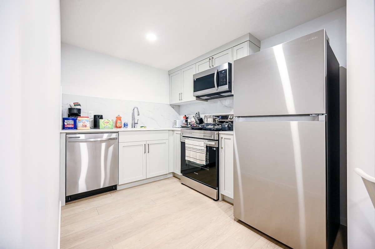 A kitchen area is presented, featuring modern stainless-steel appliances, including a refrigerator, stove, and dishwasher. Light-colored cabinetry complements the white marble backsplash, while the countertop provides ample workspace. Natural light illuminates the room through overhead lighting.