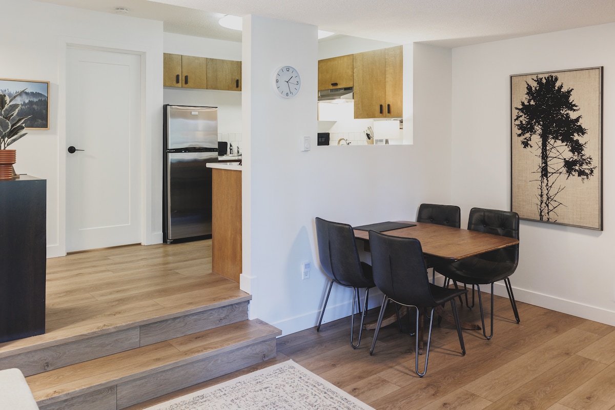 A modern dining area is visible with a wooden table surrounded by six black chairs. The open kitchen features wooden cabinetry and stainless-steel appliances. Light wooden flooring and a wall clock enhance the contemporary feel of the space, while minimalistic art adorns one wall.