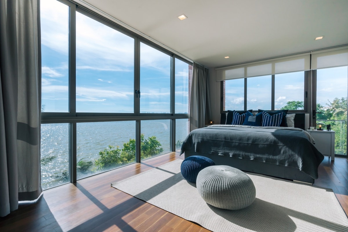A spacious bedroom features large floor-to-ceiling windows, offering expansive views of the ocean. The bed is adorned with decorative pillows and complemented by a cozy rug. A pair of textured poufs add a relaxed touch to the serene atmosphere.