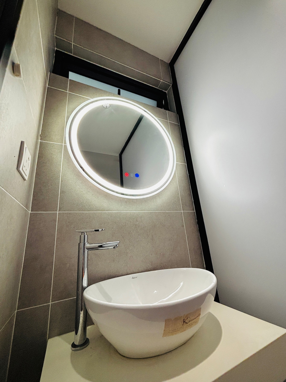 A modern bathroom features a circular illuminated mirror above a sleek basin. The walls are tiled in neutral tones, and natural light is softly filtered through a nearby window, enhancing the contemporary design.