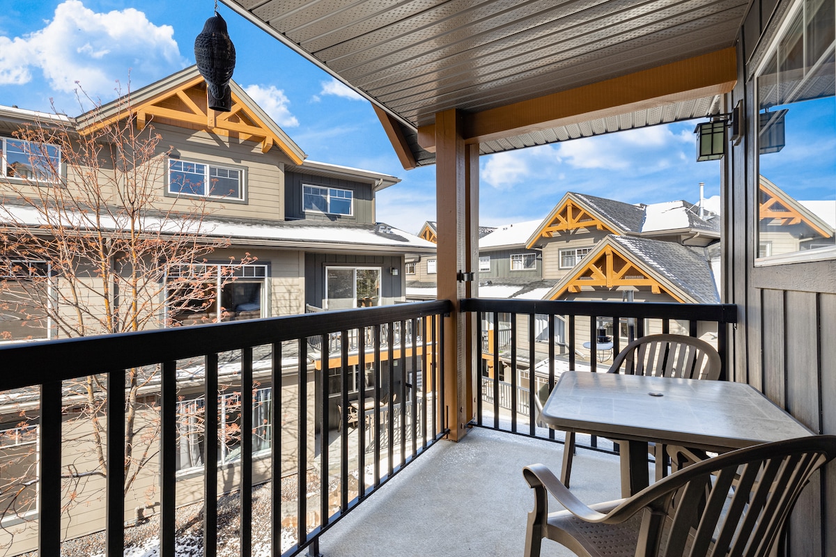 The private balcony features a small table and two chairs, providing a serene spot to enjoy the views of nearby buildings. The structure is complemented by wooden accents, with sunlight illuminating the patio area under a clear blue sky.