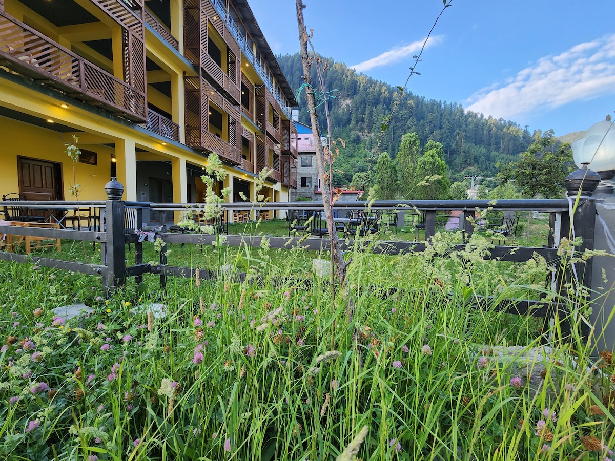 A lush garden area features tall grasses and colorful wildflowers, framed by a rustic wooden fence. In the background, the structure displays multiple balconies and large windows, surrounded by green trees and mountainous terrain under a clear blue sky, offering a serene setting.