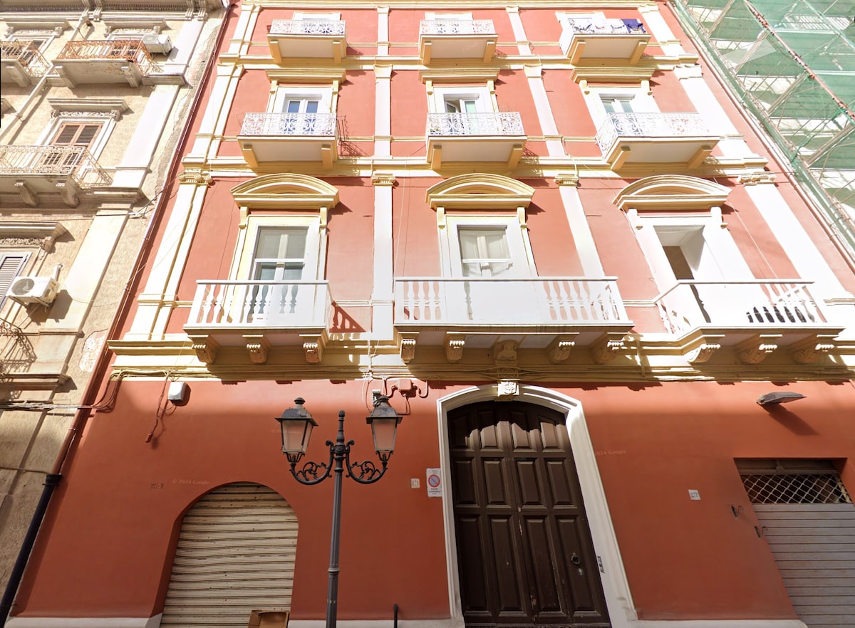A historic building is featured, displaying a charming facade in warm tones. Balconies with ornate railings can be seen above a large wooden entrance door, flanked by decorative lamp posts. The surrounding area includes architecture with a blend of colors and textures.