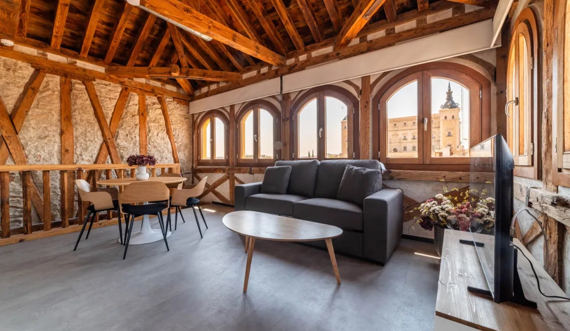A spacious living area features large arched windows, providing views of the surrounding area. A comfortable sofa is positioned across from a television. A dining table with chairs occupies one corner, and decorative elements like dried flowers add character to the space.