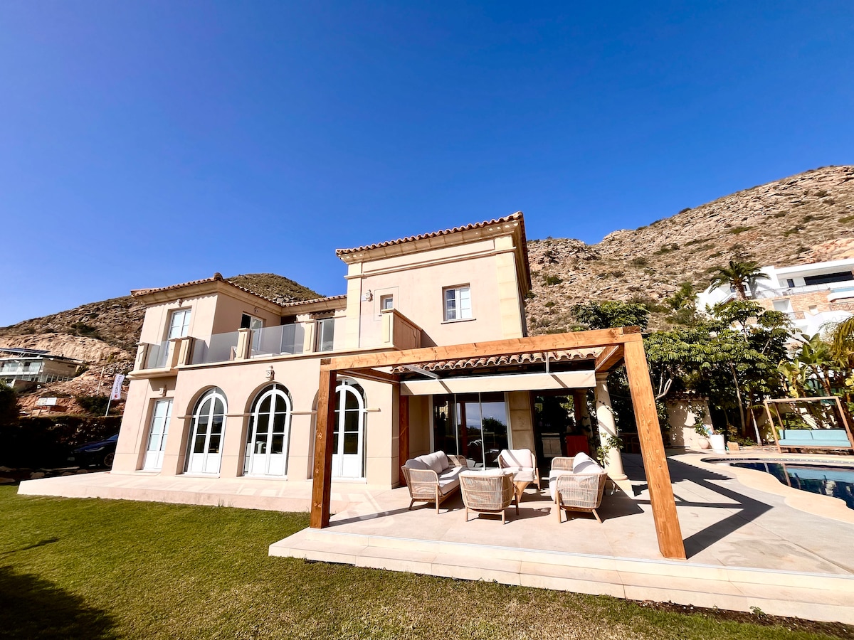 A spacious villa is presented with a light-colored exterior, set against a mountainous backdrop. A wooden pergola provides a shaded seating area, accompanied by four comfortable chairs. Large arched windows enhance the architecture, while a well-maintained lawn complements the outdoor setting.