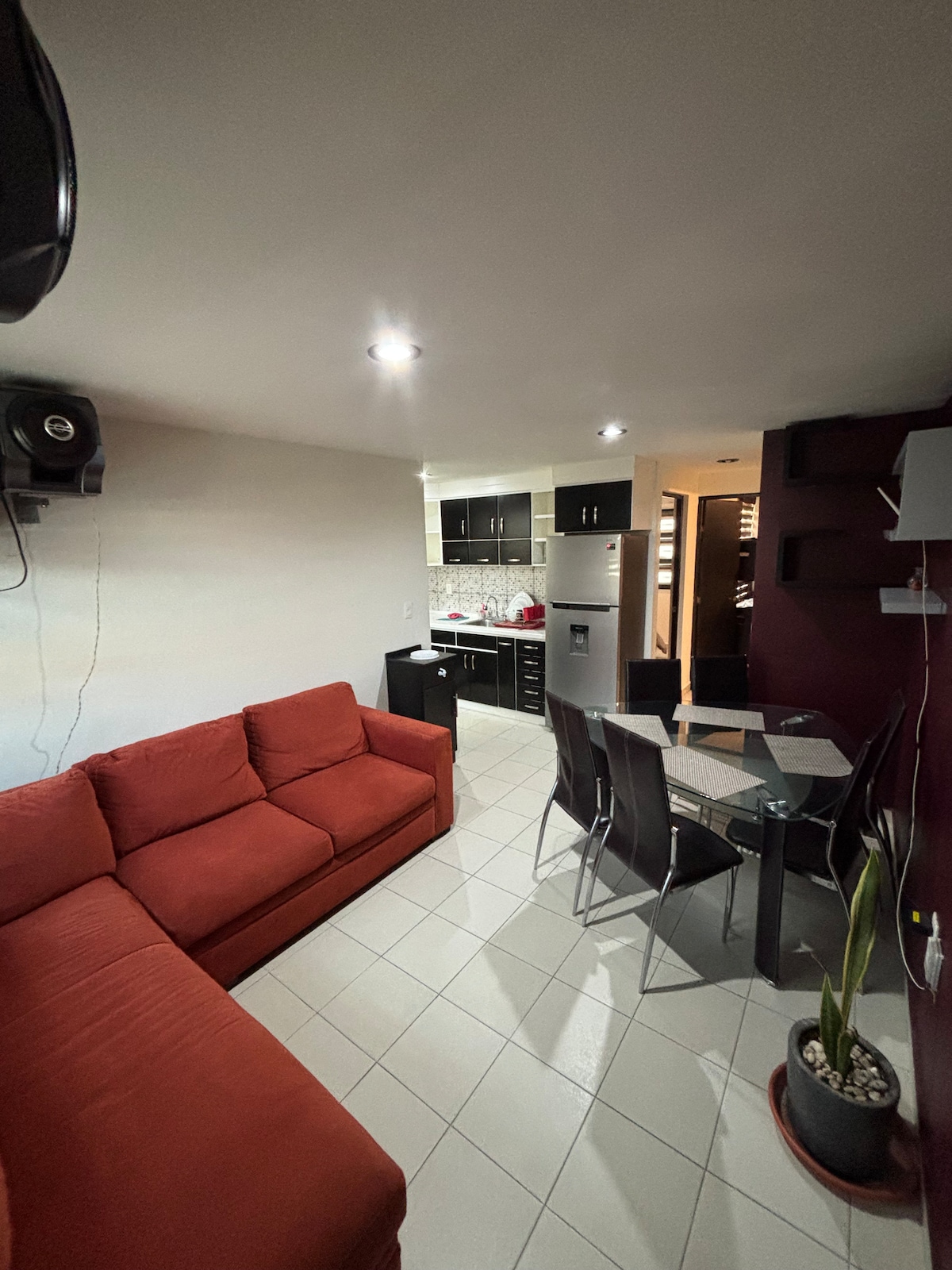 A modern living area features a red sectional sofa and a glass dining table surrounded by four black chairs. Soft overhead lights illuminate the space, while a glimpse of the kitchen with dark cabinetry and light countertops can be seen in the background.