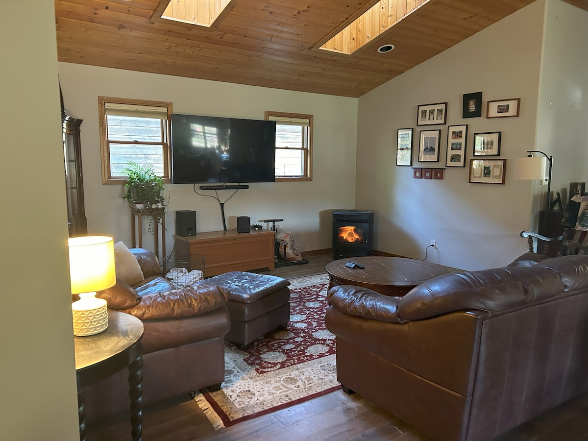 A comfortably arranged living area features a large leather sectional sofa and an ottoman. A television is mounted on the wall, above a wooden media console. A warm fire flickers in a corner fireplace, and framed photographs adorn the walls. Natural light filters through windows.