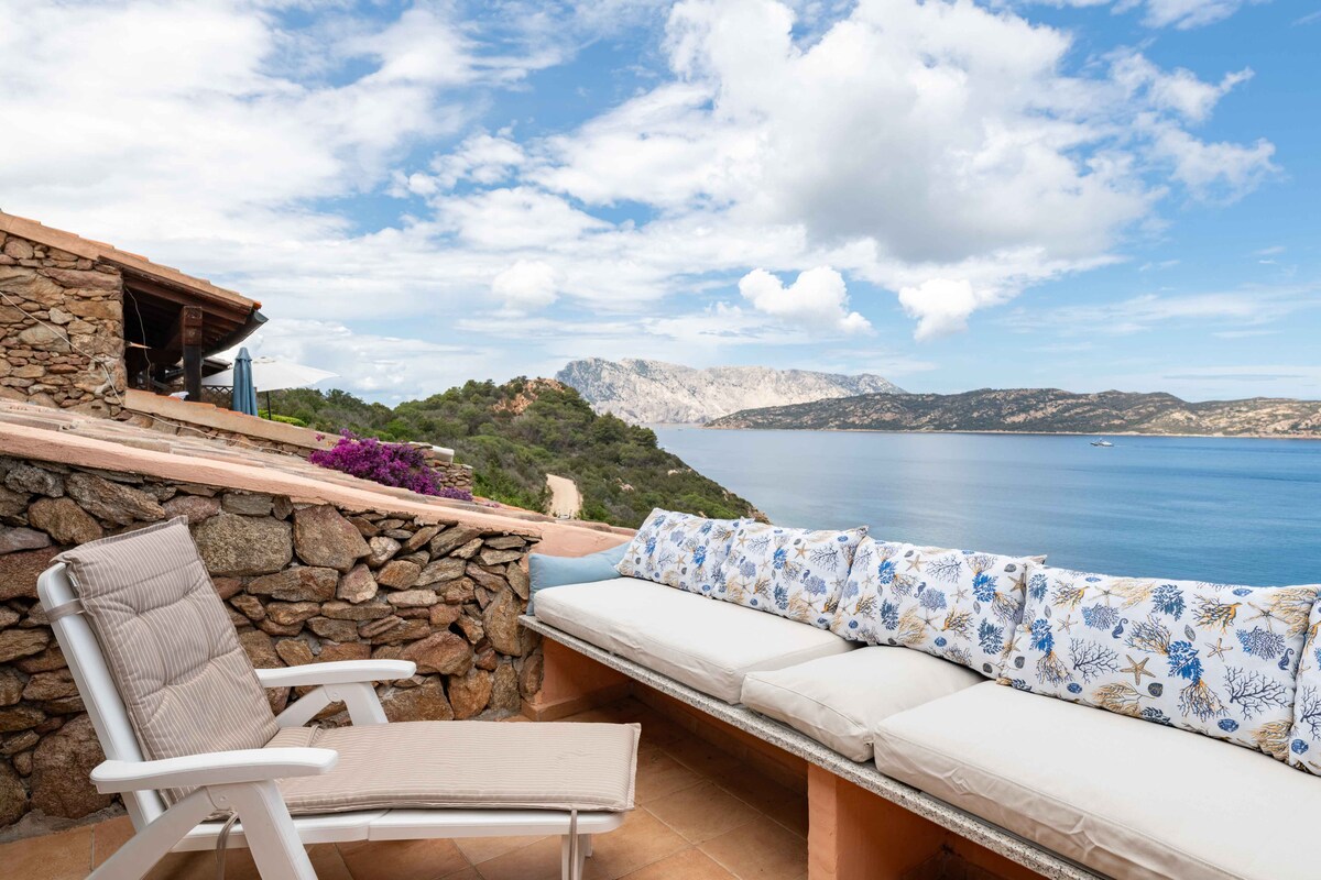 A spacious terrace features a comfortable seating area with a textured cushion along the wall, overlooking the serene sea and the distant islands. A single lounge chair is positioned nearby, framed by natural stone walls and lush greenery.