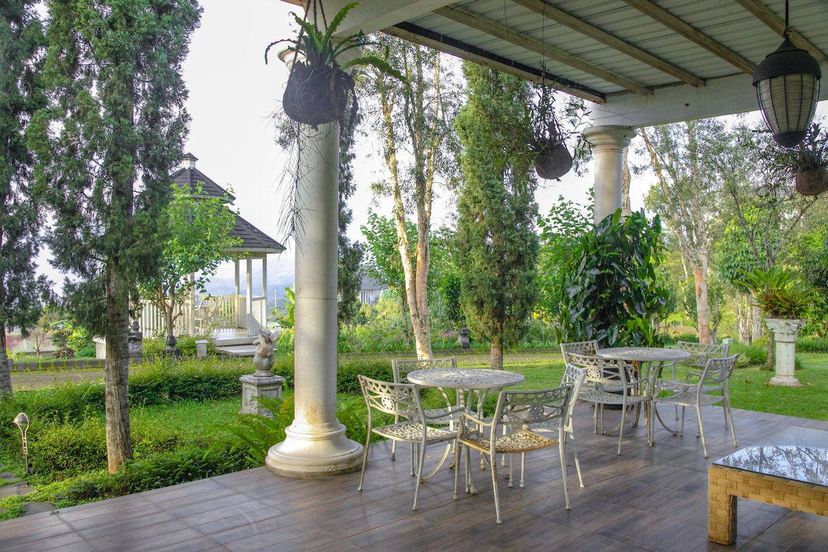 A shaded outdoor seating area is surrounded by lush greenery, featuring a round table accompanied by six chairs. Decorative plants hang from the ceiling, and a gazebo is visible in the background, contributing to the peaceful atmosphere of the space.