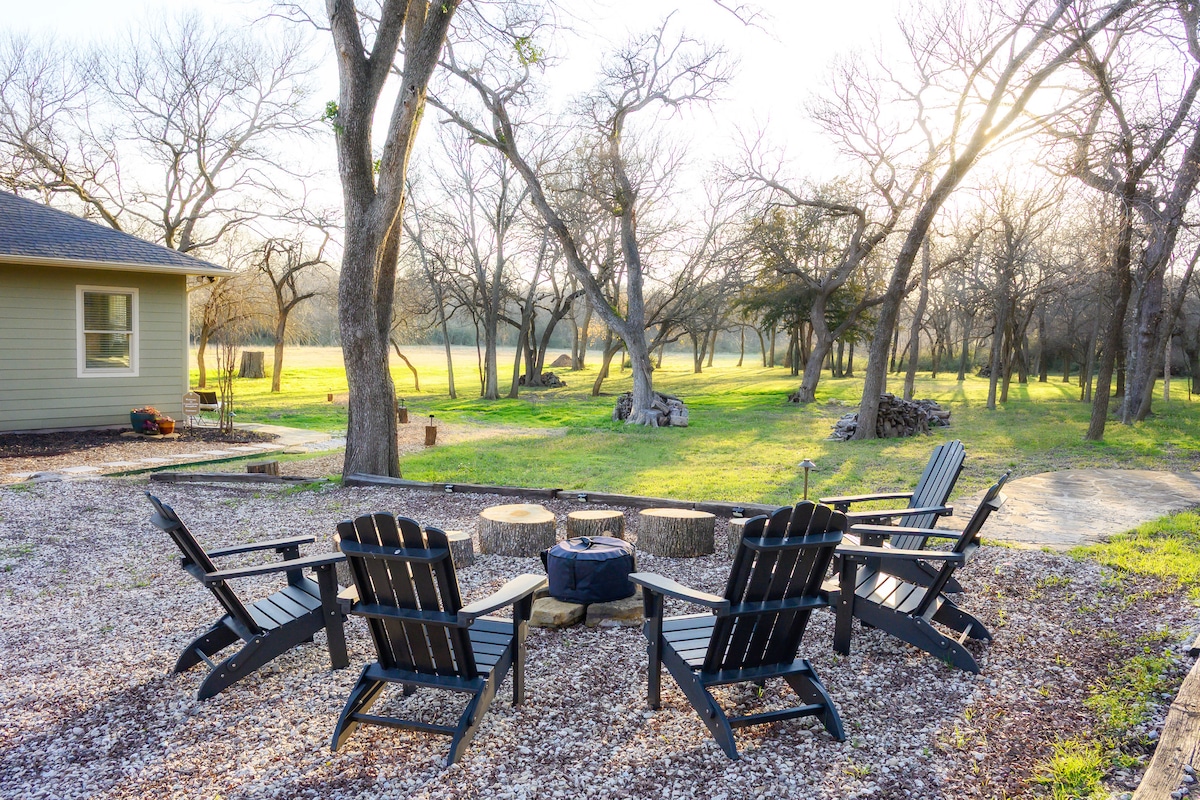 A serene outdoor seating area features several black adirondack chairs arranged around a central firepit, surrounded by gravel. The background showcases a sprawling green lawn bordered by bare trees, with soft sunlight filtering through, creating a tranquil atmosphere.