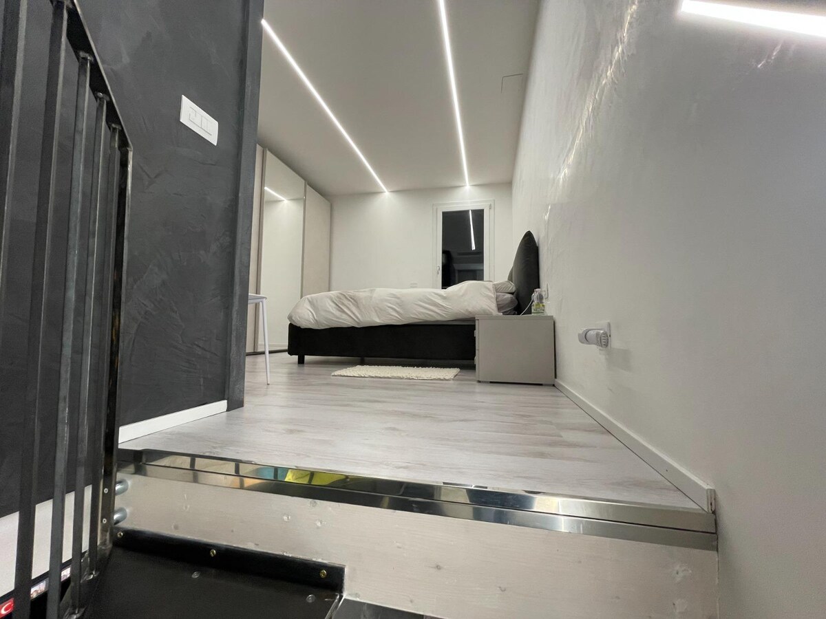 The upstairs sleeping area features a spacious bed with a minimalistic design. Soft lighting is provided by recessed fixtures along the ceiling, and a large window offers natural light. The floor is sleek and modern, complementing the overall aesthetic of the apartment.