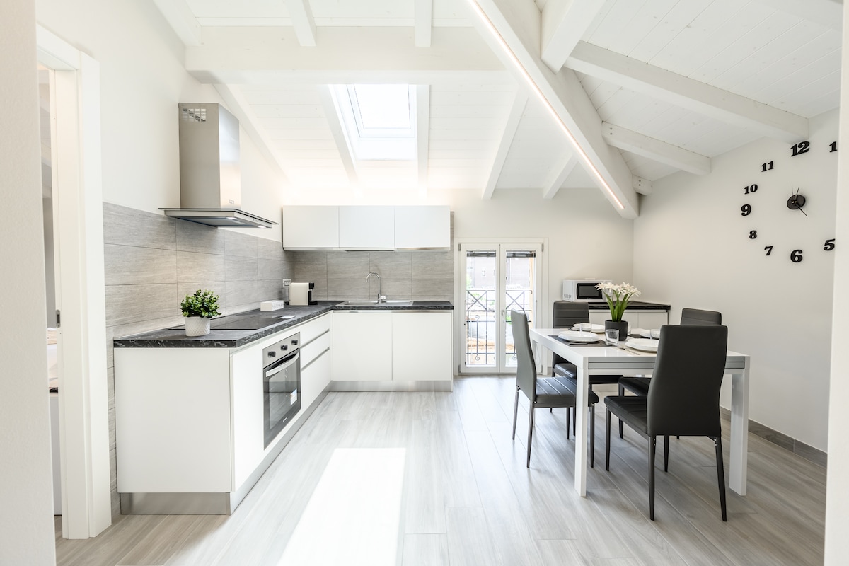 A modern kitchen and dining area features white cabinetry and a granite countertop. Large windows allow natural light to fill the space, highlighting a dining table with four chairs. A wall clock and a potted plant add a touch of detail to the bright, open design.