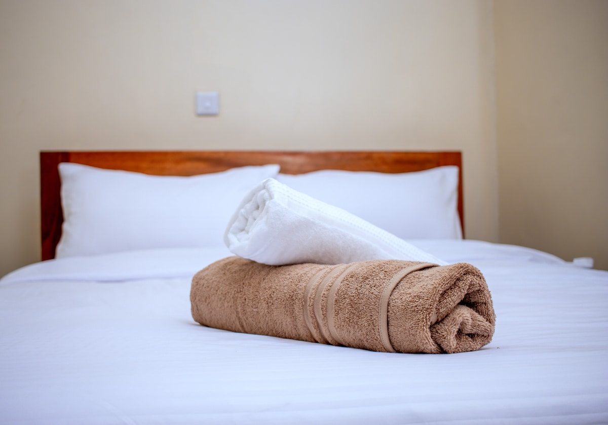 A spacious bed is neatly made with white linens, featuring two plump pillows. A rolled white towel and a rolled brown towel are placed on top, adding a touch of warmth to the arrangement.