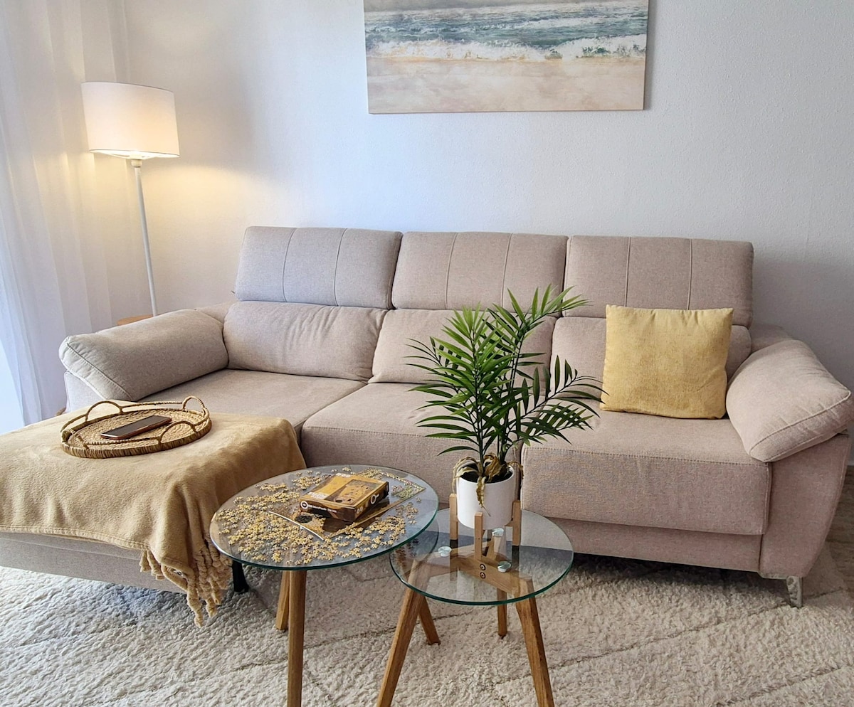 A light-filled living area features a comfortable beige sofa with an accent pillow and a soft throw draped over the arm. Two glass tables are placed in front, one adorned with a decorative tray and a small potted plant, contributing to a relaxed ambiance.