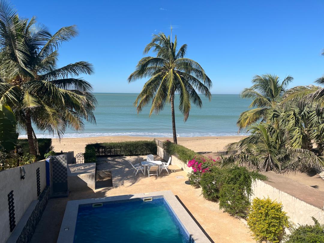 A sunlit outdoor area features a clear swimming pool, surrounded by tropical greenery. Palm trees frame the scene, leading to a sandy beach and calm sea in the background. Loungers are positioned nearby, creating a serene setting for relaxation.