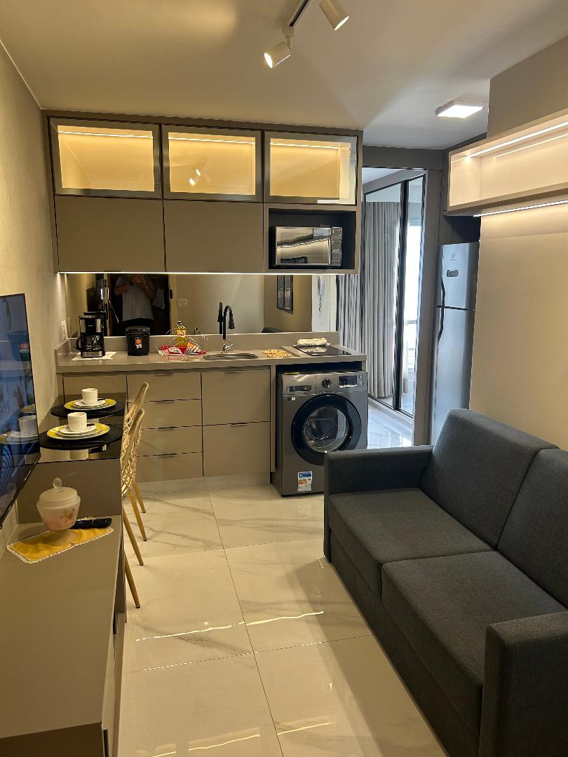 A modern kitchen area with sleek cabinetry features a compact dining setup for two. Appliances include a washing machine, coffee maker, and refrigerator. Natural light enters through a large window, adding brightness to the space. The sofa provides additional seating within the studio layout.