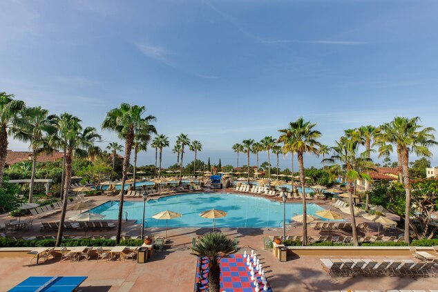 A spacious pool area is framed by palm trees, featuring an expansive blue pool surrounded by sun loungers and umbrellas. Various seating options are scattered along the deck, creating a welcoming environment for relaxation.