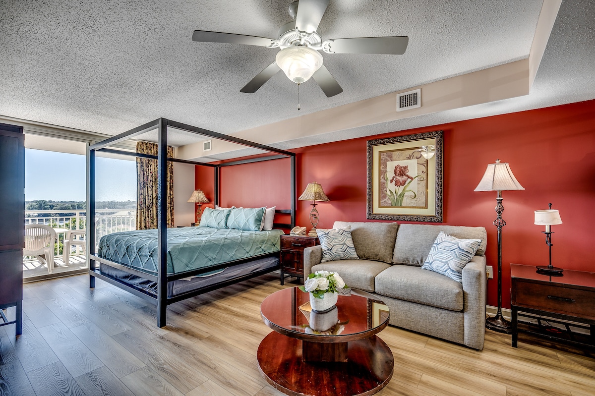 A spacious studio is highlighted by a four-poster bed draped in blue bedding, complemented by a light gray sofa. Two lamps provide ambient lighting, and a round coffee table sits in the center. A sliding door offers access to a balcony with views of the waterway.