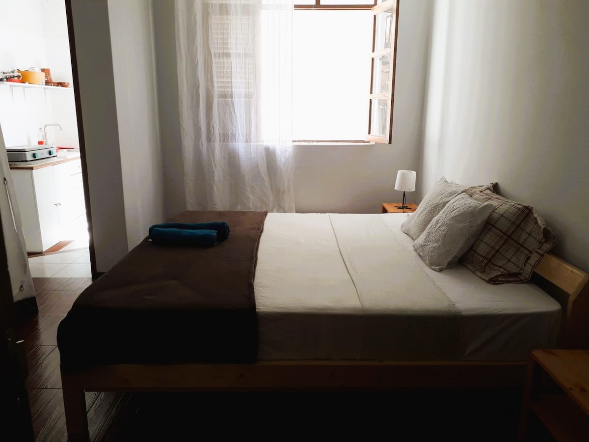 A spacious bed is prominently featured, dressed in neutral bedding with a dark throw and a rolled towel on top. A window covered with sheer curtains allows natural light to fill the room, complementing the wooden furniture and bedside lamp.