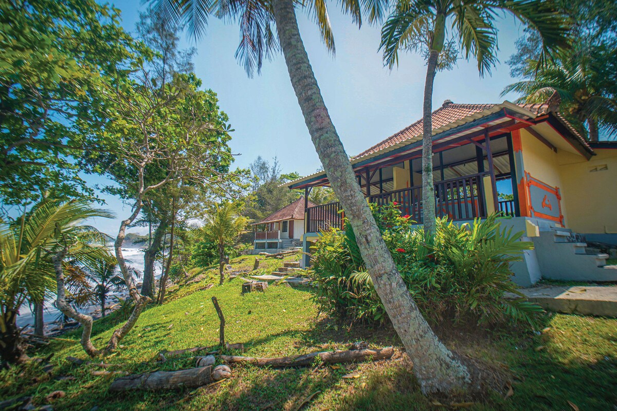 A charming beachfront property is situated amidst lush greenery, featuring palm trees and vibrant foliage. The structure showcases a covered patio with wooden railings and a terracotta-tiled roof, offering peaceful views of the ocean and surrounding nature.
