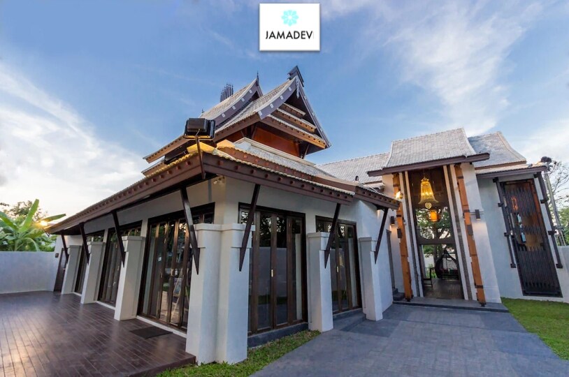 The exterior of Jamadevi hotel is depicted, showcasing a blend of traditional and modern architectural elements. The sloped roof features decorative details, and large glass windows invite natural light. A spacious wooden deck leads to the entrance, framed by lush greenery in the surroundings.