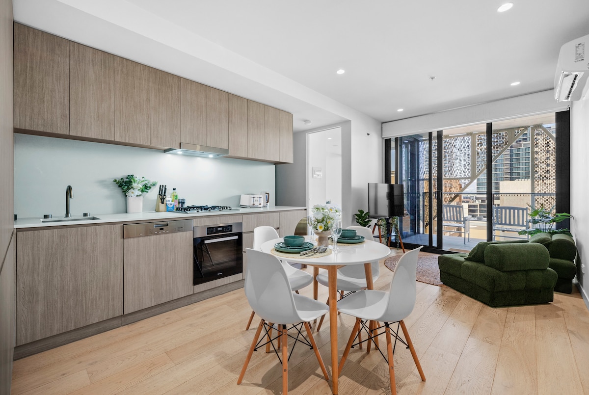 A modern kitchen area with wooden cabinetry and stainless steel appliances is depicted. A round dining table with white chairs is set for four. Floor-to-ceiling glass doors open to a balcony, allowing natural light to fill the open living space, which features a cozy green couch.