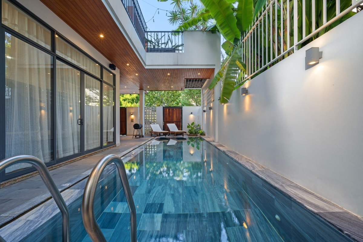 A private pool is framed by a sleek stone deck, with lounge chairs positioned nearby. Sunlight reflects off the water's surface, while greenery adds a tranquil touch. Large glass doors open to the interior, creating a seamless connection between indoor and outdoor spaces.