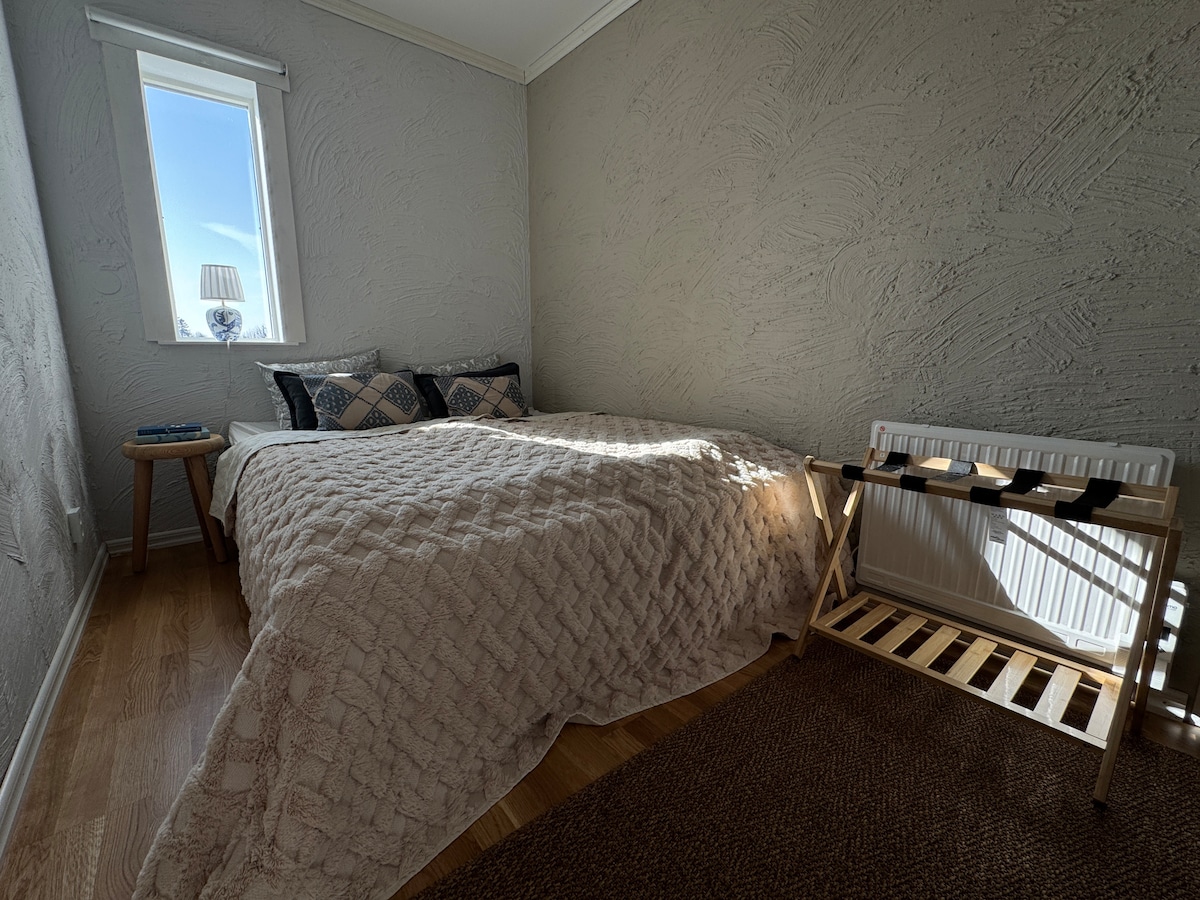 A cozy bedroom features a spacious bed adorned with a light-colored quilt and decorative pillows. Natural light filters through a small window, illuminating the room. A wooden stool and a drying rack are positioned beside the bed, contributing to the functional layout.