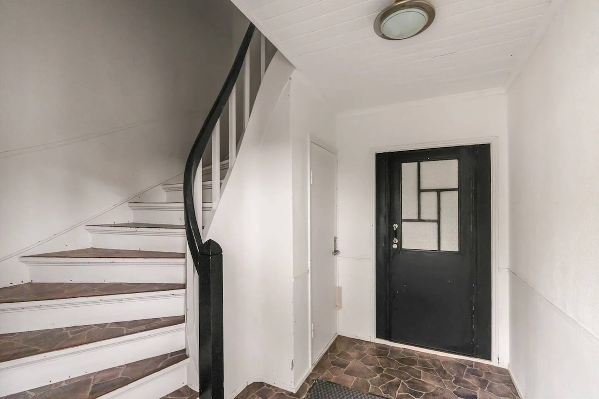 The entrance features a staircase leading to the upper level, complemented by a simple light fixture overhead. The floor is made of textured tiles, and a black door with geometric glass accents serves as the main entry point.