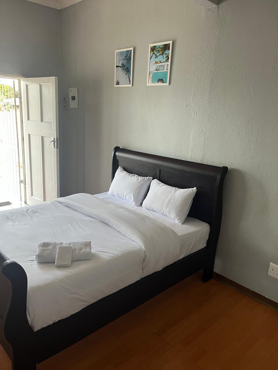 A bright sleeping area features a dark wood bed with a white duvet and two white pillows. Towels are neatly arranged at the foot of the bed. Natural light enters through an open door, enhancing the calming atmosphere, complemented by two framed photographs on the wall.