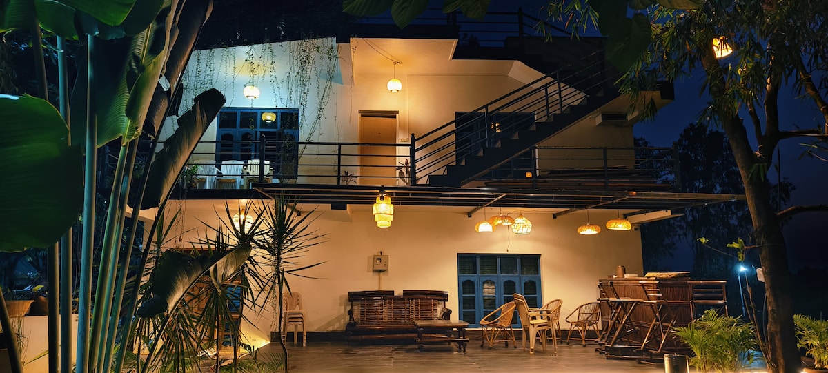 The exterior of a multi-level building is illuminated at night, showcasing a harmonious blend of natural and architectural elements. Various light fixtures hang from the ceiling, highlighting outdoor seating areas which include wooden chairs and tables. Lush greenery surrounds the space, adding to the tranquil ambiance.