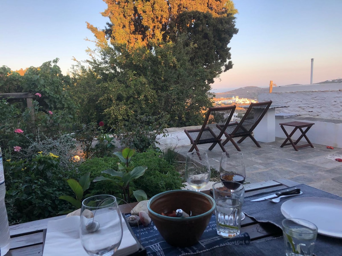 An outdoor dining area is visible, featuring a table set with glasses and a bowl. Two wooden chairs are situated nearby, surrounded by lush greenery and blooming flowers. A serene landscape is in the background, showcasing the hills and distant buildings under a soft sunset light.